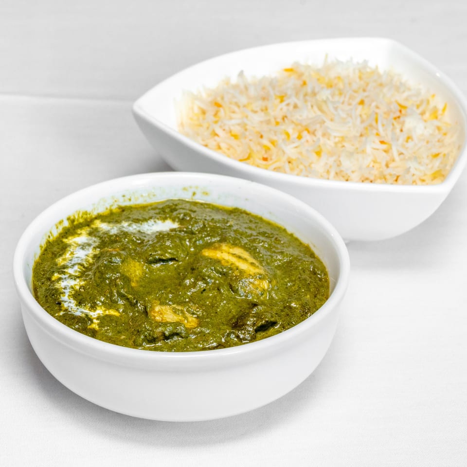 Bowl of creamy green spinach curry with paneer alongside a dish of fragrant rice on a white table.