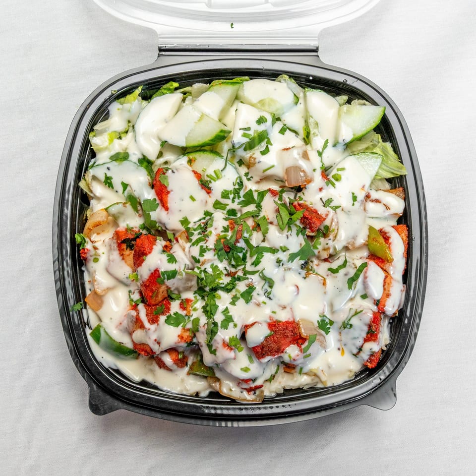 Takeout container filled with creamy-dressed salad, grilled vegetables, red-seasoned protein, cucumbers, lettuce, and chopped herbs.