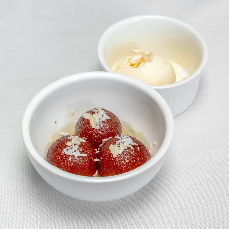 Three red gulab jamun desserts topped with coconut and almonds sit in a white bowl, with vanilla ice cream in a second bowl behind.