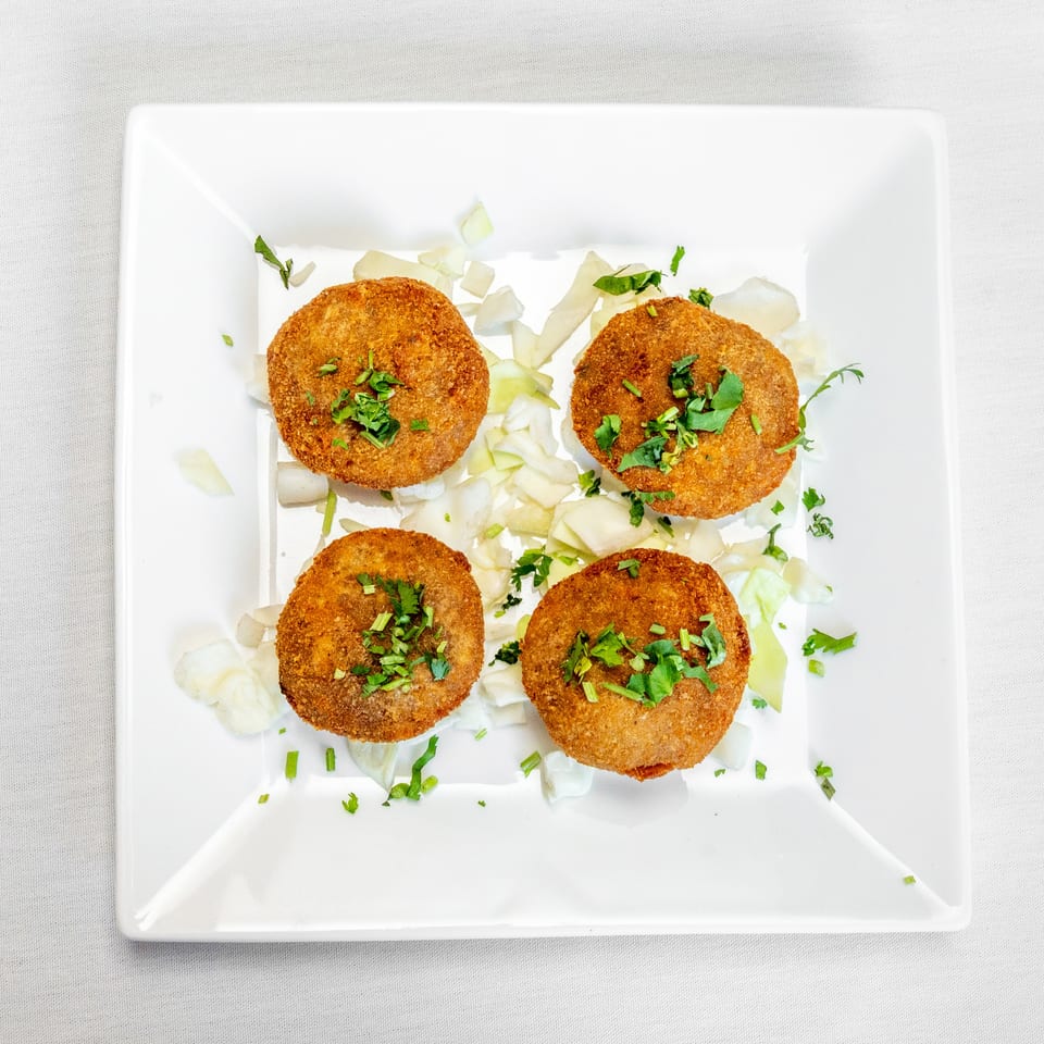 Four golden-brown breaded patties garnished with chopped herbs and onions on a square white plate.
